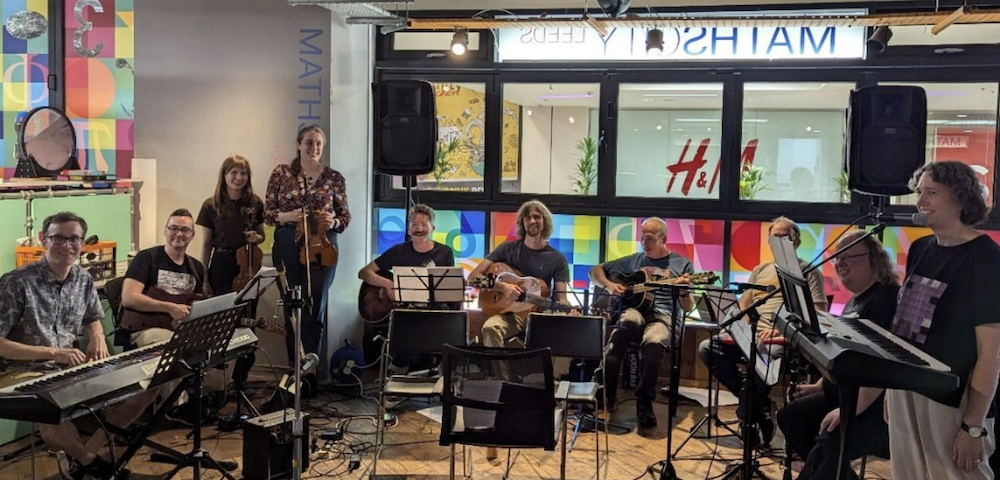 Photo of musicians in MathsCity, seated in a line against a window through which a shopping centre is visible